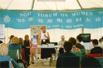 [ai] A woman speaking at a podium during the NGO Forum on Women '95 in a tent. Attendees are seated with papers in front of them. A blue banner displays event details and location.